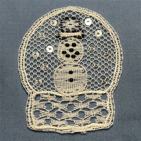 A lace applique depicting a snowman, featuring cream-colored mesh embroidery with black details and small silver sequins, set against a textured blue-gray fabric background.